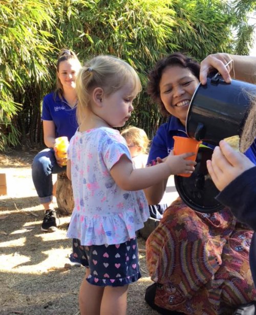 Educator crouching down outside pouring water into a young child's orange cup