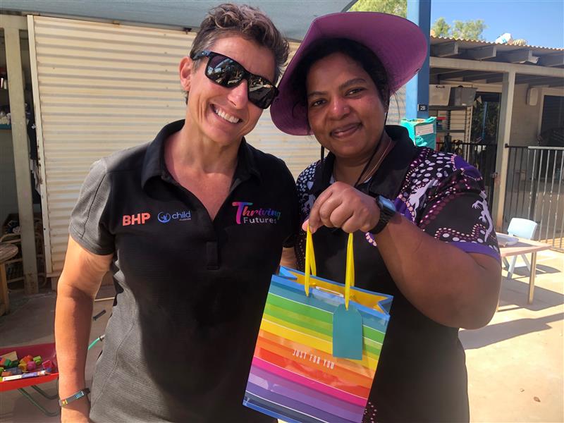 Two women smiling, one with sunglasses on, the other with a broad brimmed hat holding a rainbow coloured gift bag up to the camera