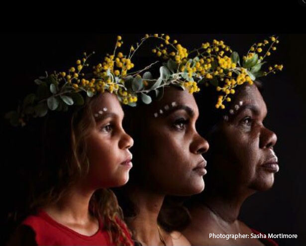 Three women of three generations against black background all facing the right, wearing native Australian yellow flower crowns with white face paint dotted above their eyebrows